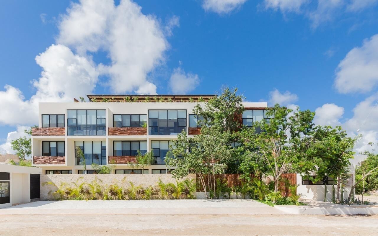 A modern multi-story building with large windows, a flat roof, and a landscaped area with trees and shrubs.