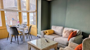 Een woonkamer met een bank, fauteuil en salontafel, een eettafel met stoelen en een raam met zonwering.