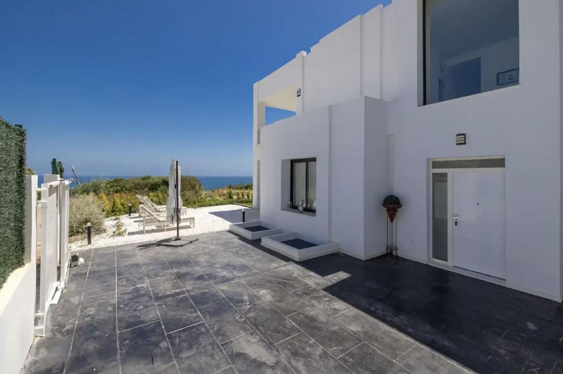 A modern building with a flat roof, a small outdoor seating area with chairs and a table, and a view of the sea.