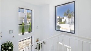 Una habitación luminosa con paredes blancas, barandilla blanca y varias ventanas que ofrecen vistas a edificios y zonas verdes.