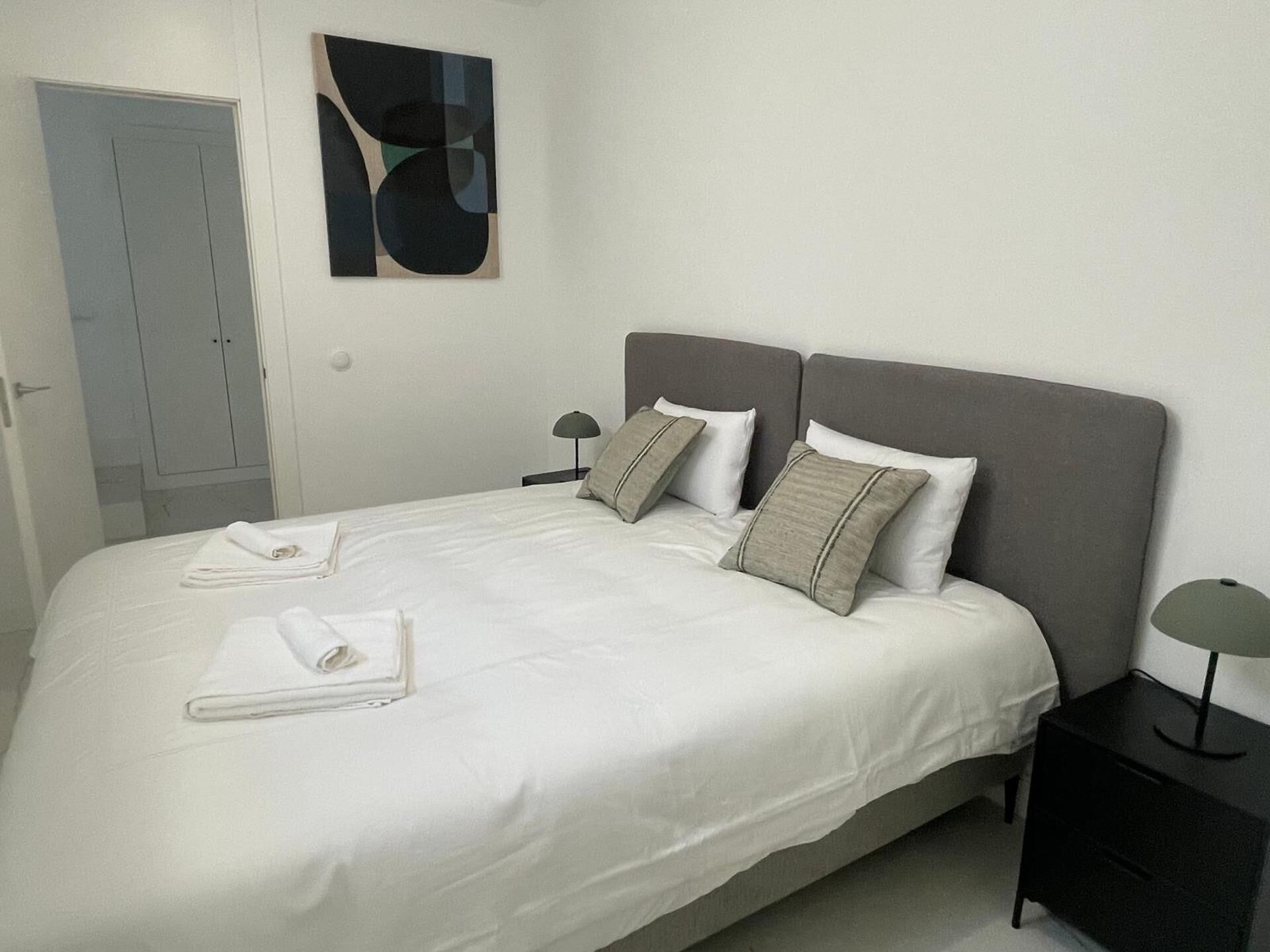 Una cama bien hecha con almohadas y toallas dobladas, una mesita de noche con lámpara y una puerta al fondo.