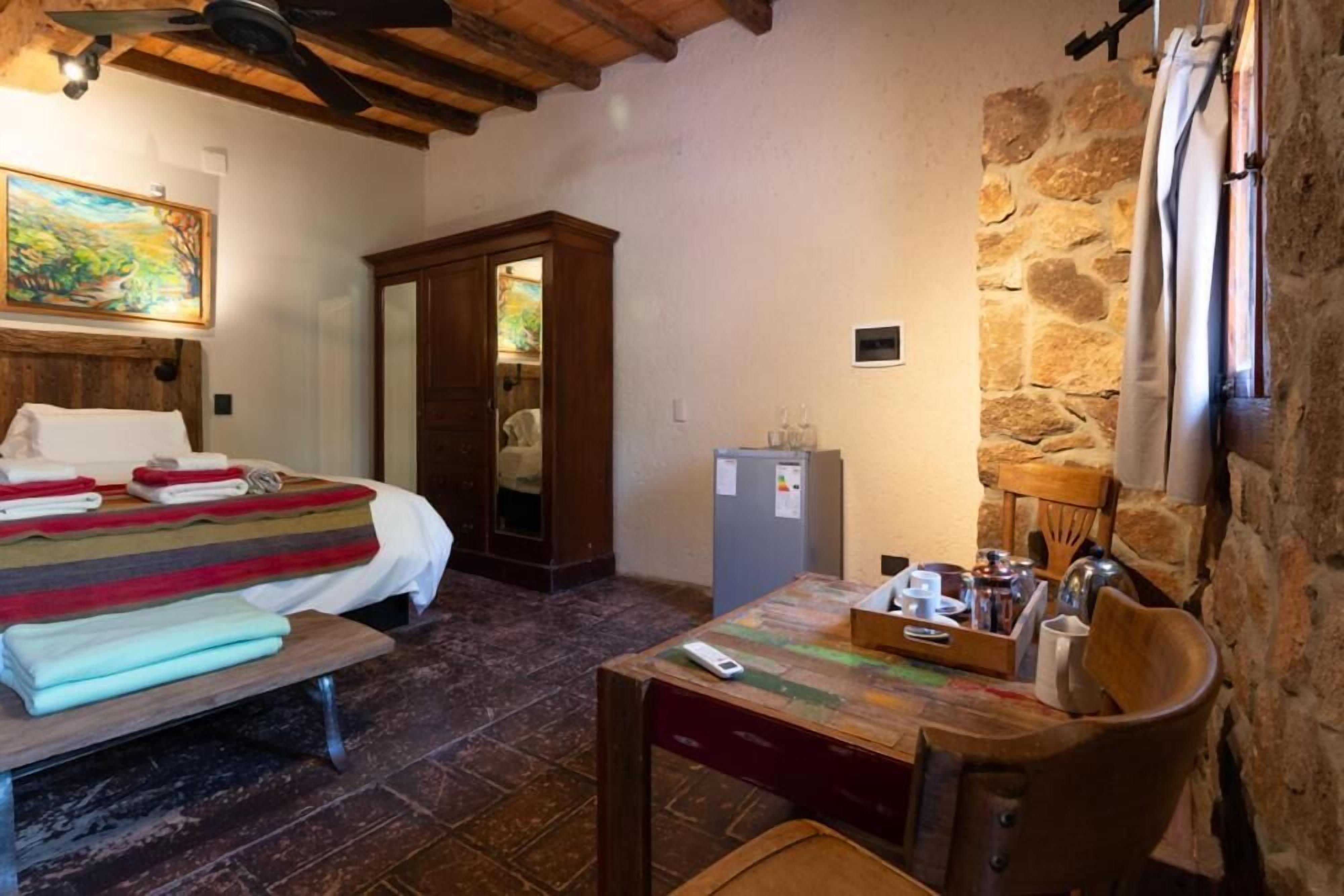 Quarto com uma cama, uma mesa de madeira com um tabuleiro, um frigorífico e uma parede de pedra.