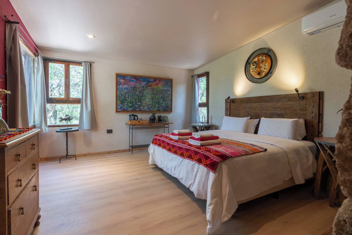 A bedroom with a wooden headboard, a bed with white linens, a colorful painting on the wall, and a wooden dresser.