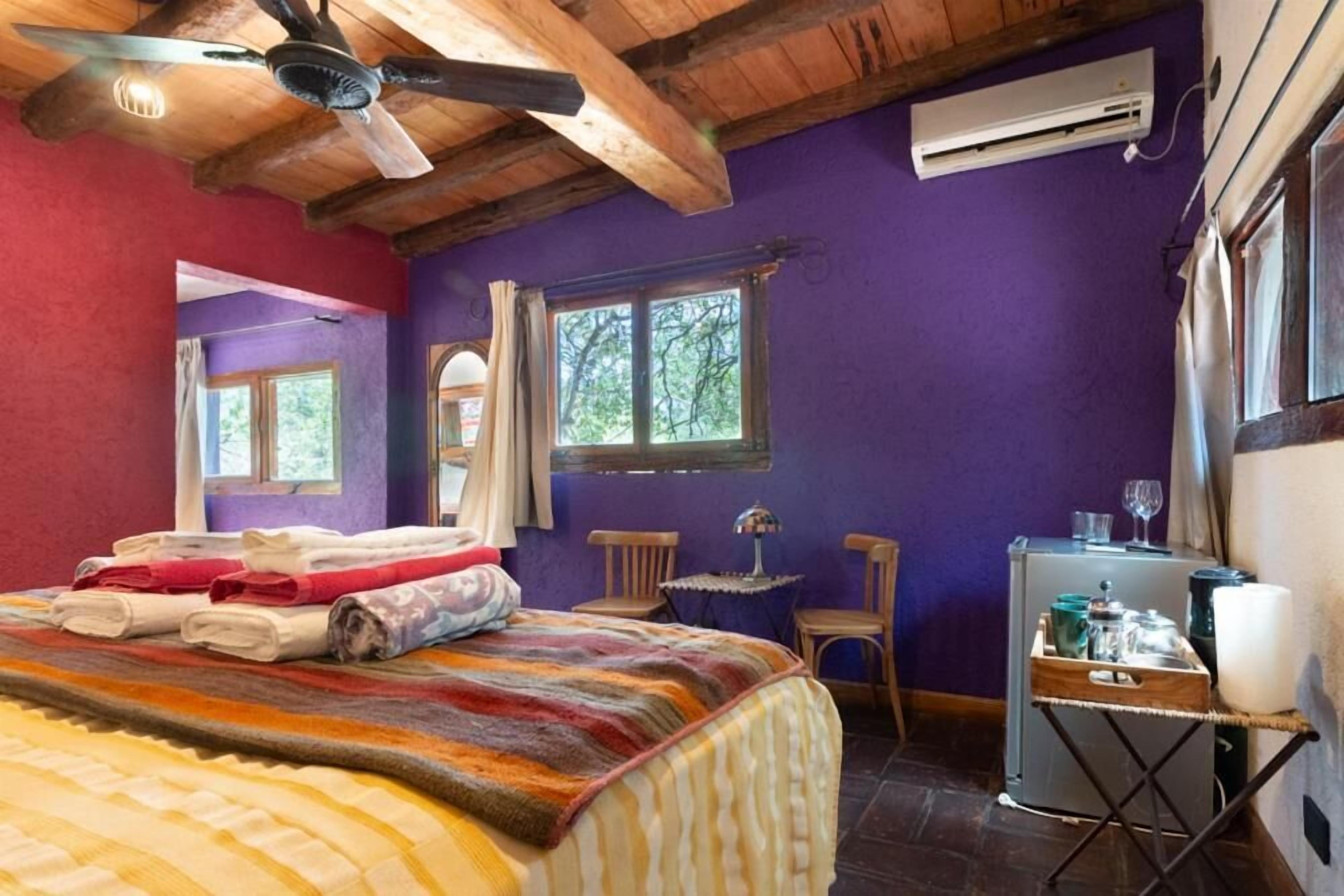 Una habitación con cama, ventilador de techo, ventana, mesa con lámpara y aire acondicionado.