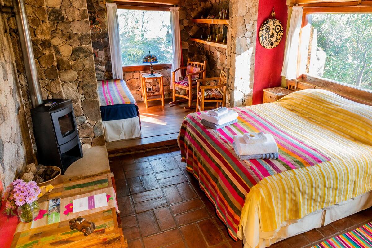 A cozy room with a bed, a small table, a chair, a fireplace, and a window.