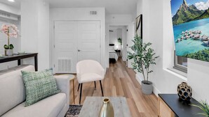 Una sala moderna con un sofá, un sillón y una mesa de centro. Hay una puerta que da a un dormitorio, una planta en maceta y un cuadro con una escena tropical.