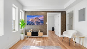 Una sala de estar moderna con un televisor grande, un sofá blanco, una pequeña mesita auxiliar, una planta en maceta y piso de madera.