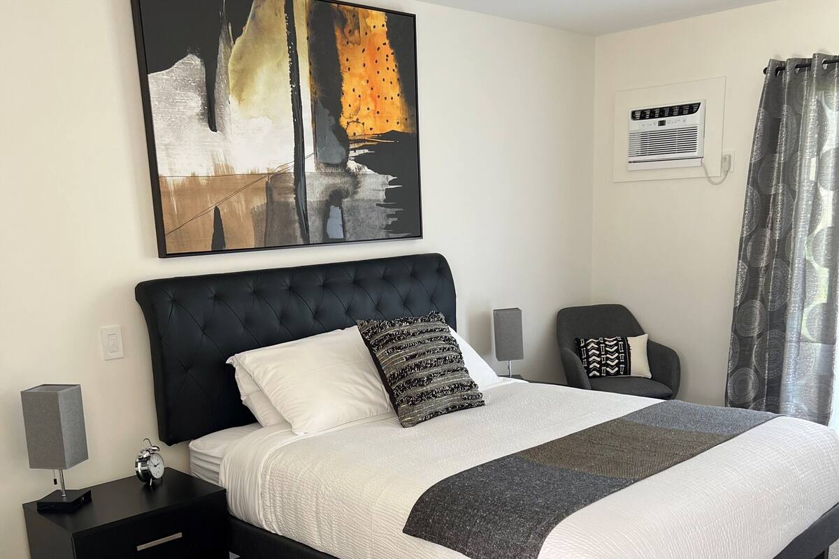 A modern hotel room with a large bed, a side table with an alarm clock, a chair, and a painting on the wall.