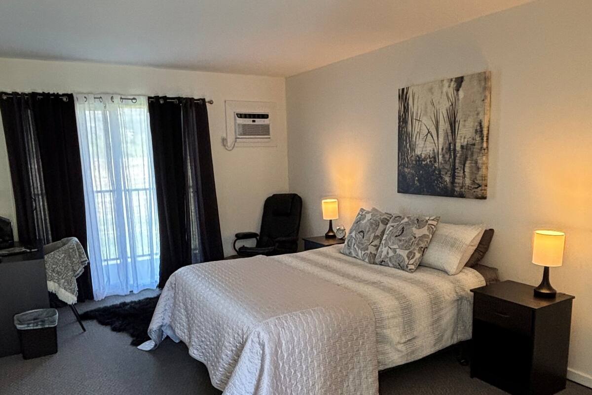 A hotel room with a bed, two bedside tables with lamps, a chair, a desk, a trash bin, and a window with curtains.