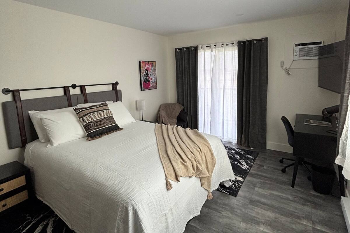 A bedroom with a bed, a desk, an air conditioning unit, and a painting on the wall.