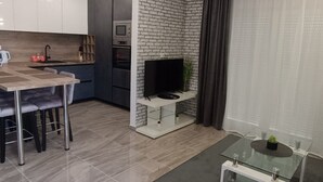 Una cocina moderna con televisor, un comedor con mesa y sillas, y una sala de estar con una mesa de centro de vidrio.