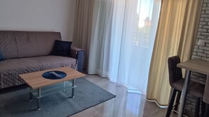 A modern living room with a sofa, a wooden coffee table, a small table with chairs, and a large window with curtains. - Dalmatia Apartments-Studio Apartment with Balcony 3a (Drage)