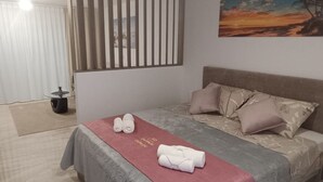 A hotel room with a bed, pillows, a red towel, a painting, a window with blinds, and a small table with a plant. - Dalmatia Apartments- Studio Apartment with Balcony and Sea View 4a (Drage)