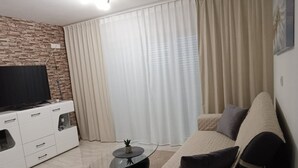 A living room with a TV on a white cabinet, a round glass coffee table, a beige sofa, and a large window with sheer curtains. - Dalmatia Apartments- Studio Apartment with Balcony and Sea View 4a (Drage)