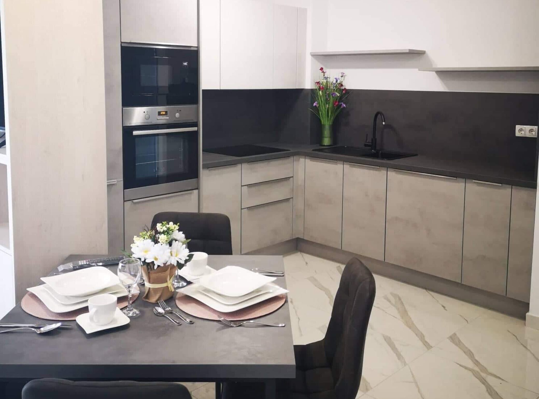 Une cuisine moderne équipée d’un four encastré, d’un évier et d’un coin repas aménagé avec une table, des chaises et un vase de fleurs.