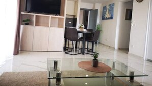 Een moderne woonkamer met een glazen salontafel, een flatscreen-televisie bevestigd aan een houten kast, een eethoek met tafel en stoelen, en een keuken op de achtergrond.