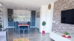 Una sala moderna con una mesa de centro de madera, un comedor con mantel rojo, una cocina con armarios blancos y una pared con textura de piedra donde está montado un televisor.