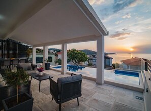 Ein überdachter Außenbereich mit Pool, Sitzgelegenheiten und Blick auf das Meer bei Sonnenuntergang.
