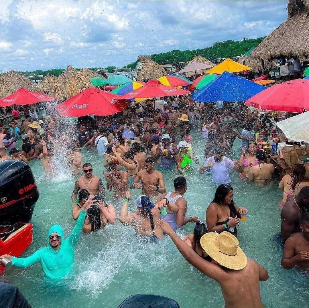 A crowded outdoor swimming area with people enjoying the water, surrounded by colorful umbrellas and thatched-roof structures.