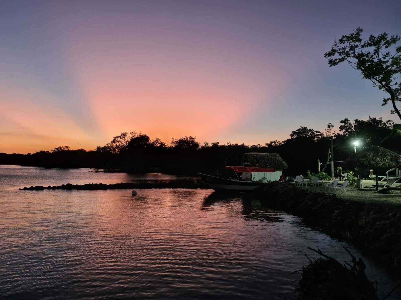 A calm water body with a boat, a thatched-roof structure, and a small building with a red roof, surrounded by trees and a clear sky.