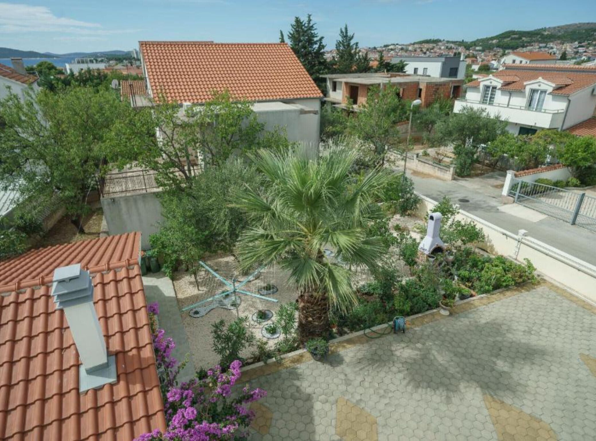 A residential area with a house featuring a tiled roof, a garden with a palm tree, and a paved driveway.