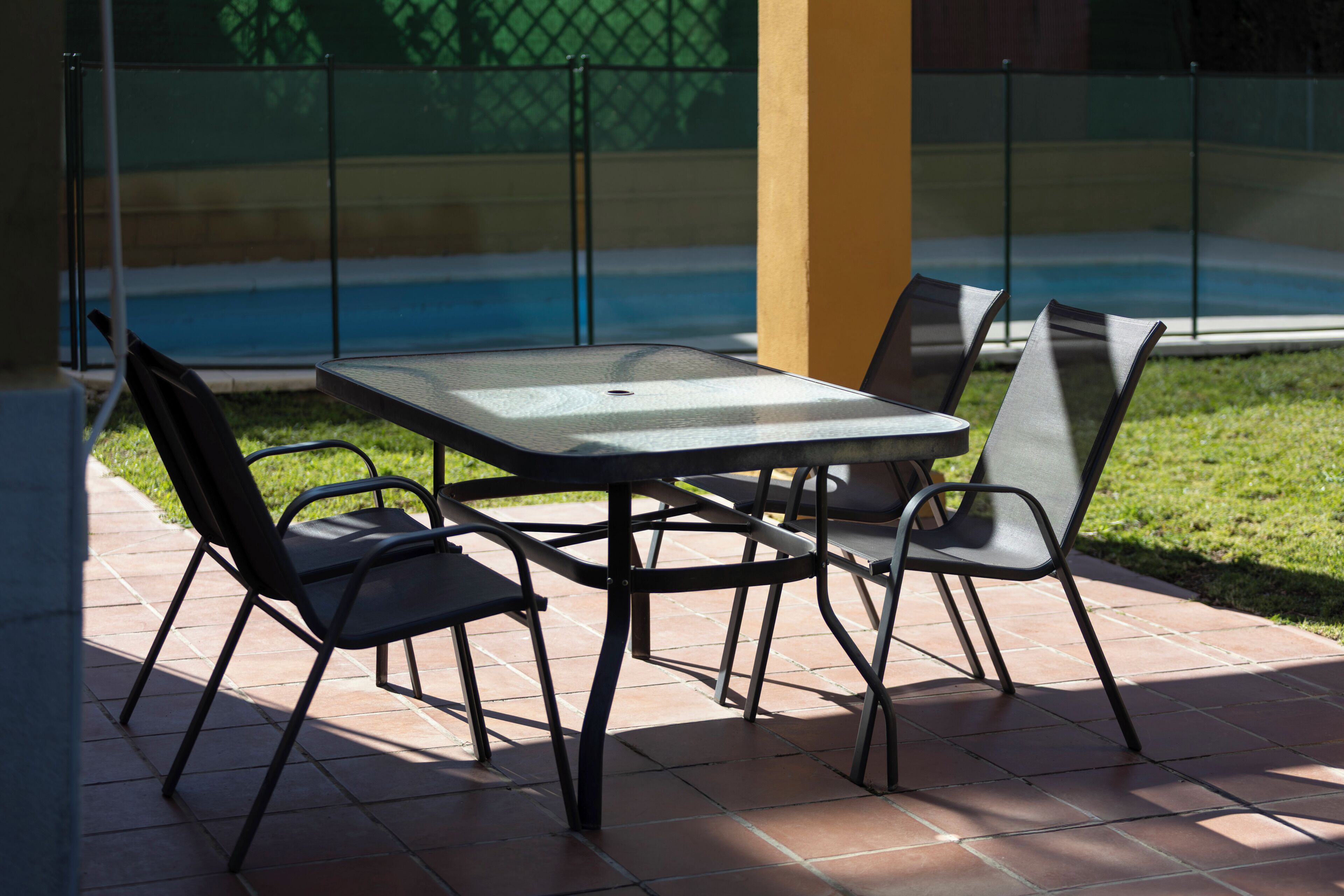 An outdoor patio with a table and chairs, a pool in the background, and a fenced area.