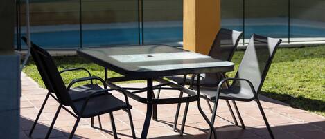 An outdoor patio with a table and chairs, a pool in the background, and a fenced area.