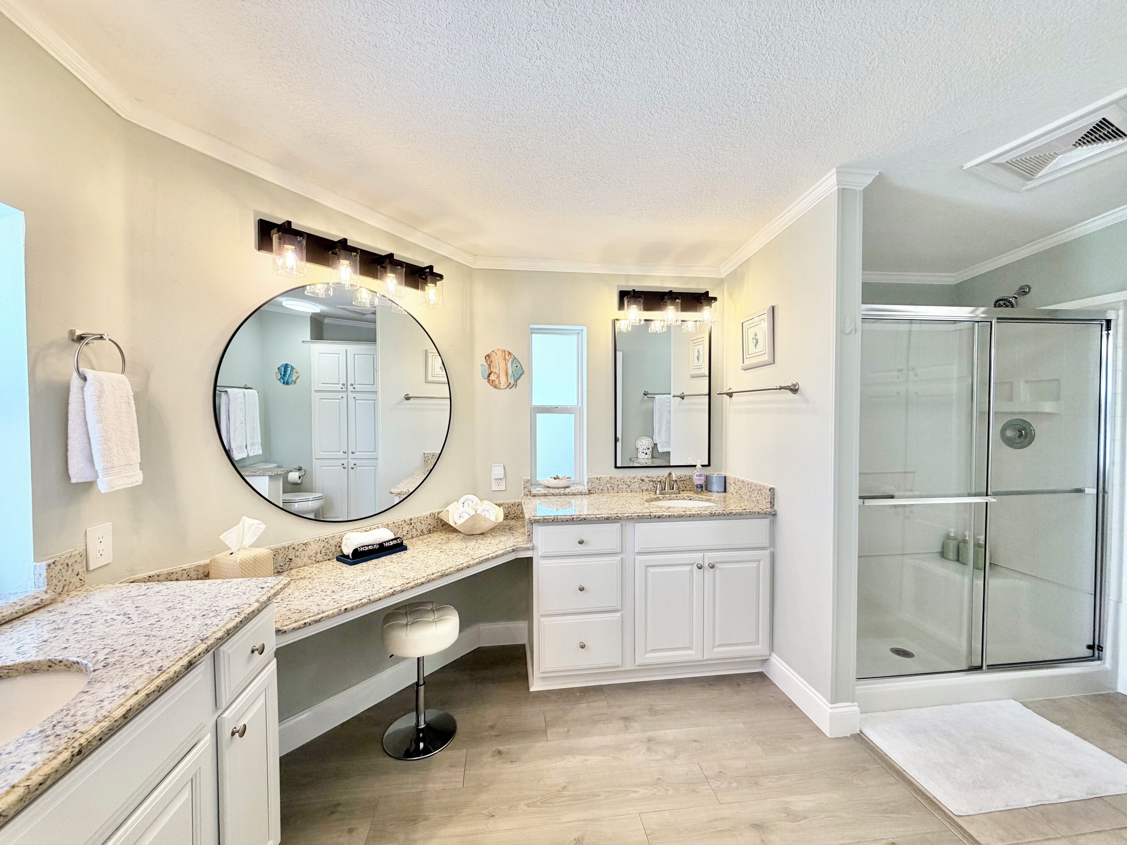 A modern bathroom with a large round mirror, a vanity with a granite countertop, a glass shower enclosure, and a wooden floor.