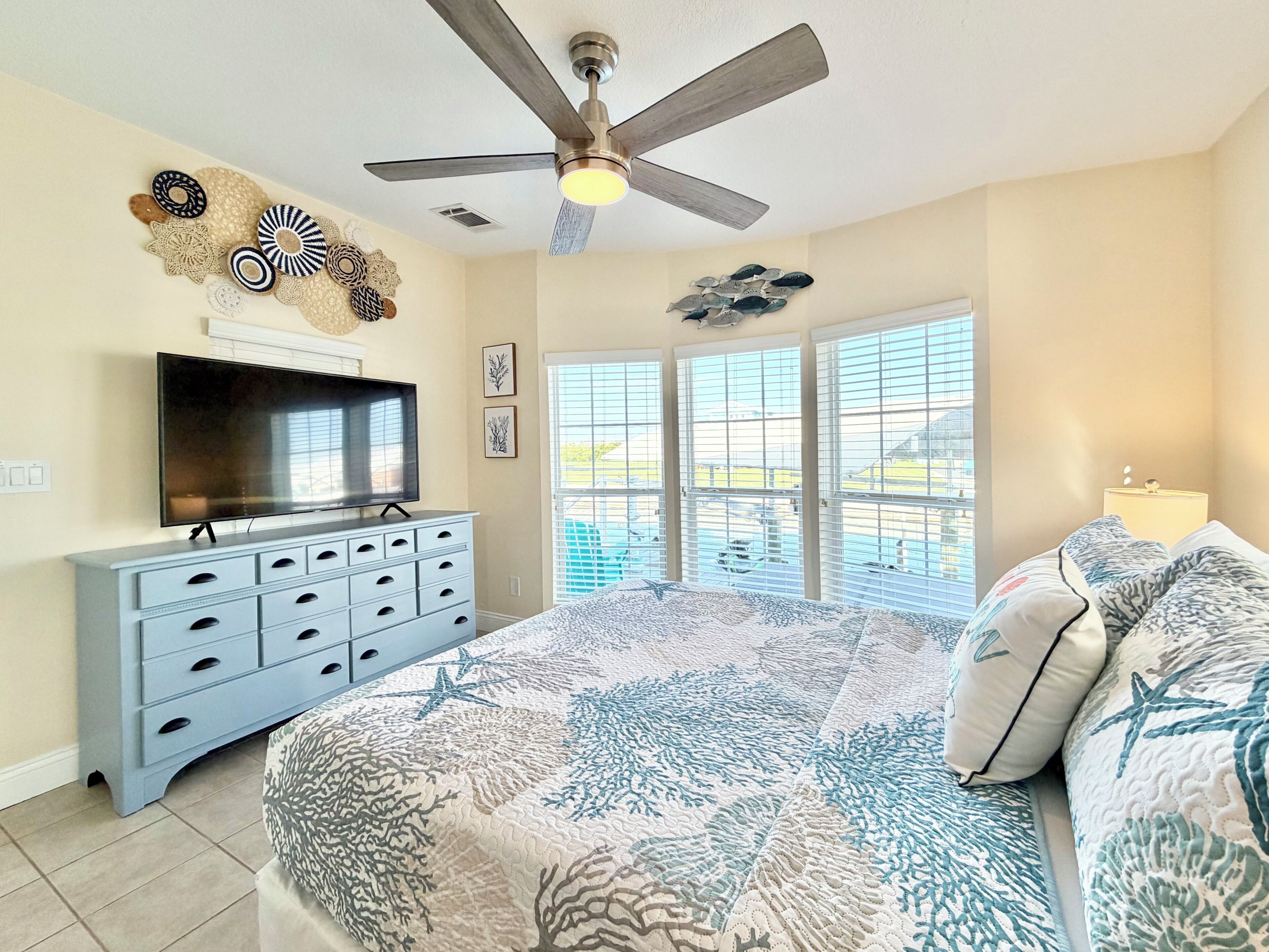 A bedroom with a large bed, a ceiling fan, a TV, and a window with blinds.