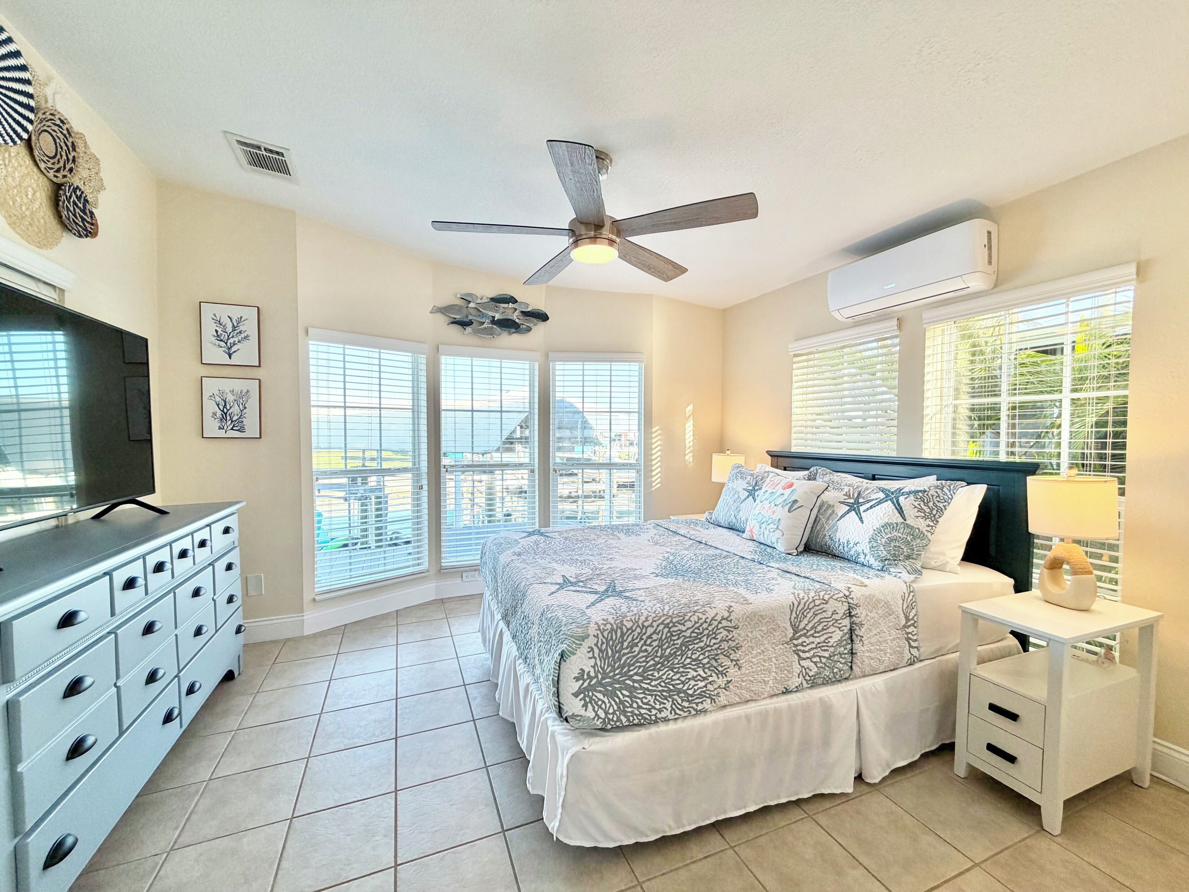 A bedroom with a bed, a ceiling fan, a TV, a dresser, and large windows.