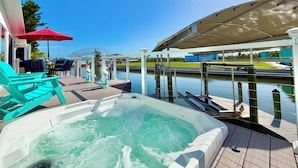A hot tub on a wooden deck with a view of a water body and a covered area with chairs and umbrellas. - Waterfront Gulf Access Home - Dock, Hot Tub, Sunset Views (Saint James City)