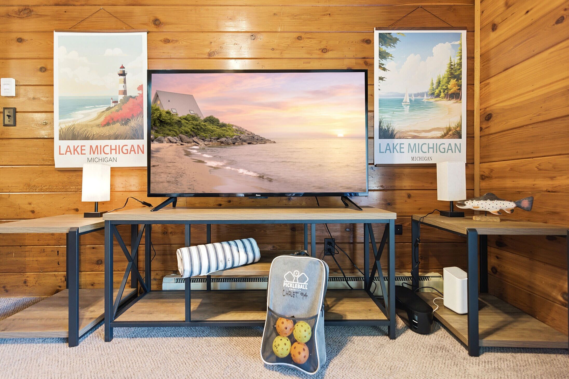 Een kamer met een televisie op een houten tafel, posters van het Michiganmeer, een pickleball tas en een vis decoratie.