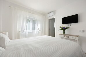 Habitación de hotel con cama, televisor montado en la pared, mesita de noche con un jarrón de flores y ventana con cortinas de tul.