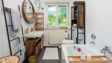 Ein modernes Badezimmer mit einem Holzbadschränkchen, einer freistehenden Badewanne, einer Toilette und einem Fenster mit Blick auf Grünflächen.