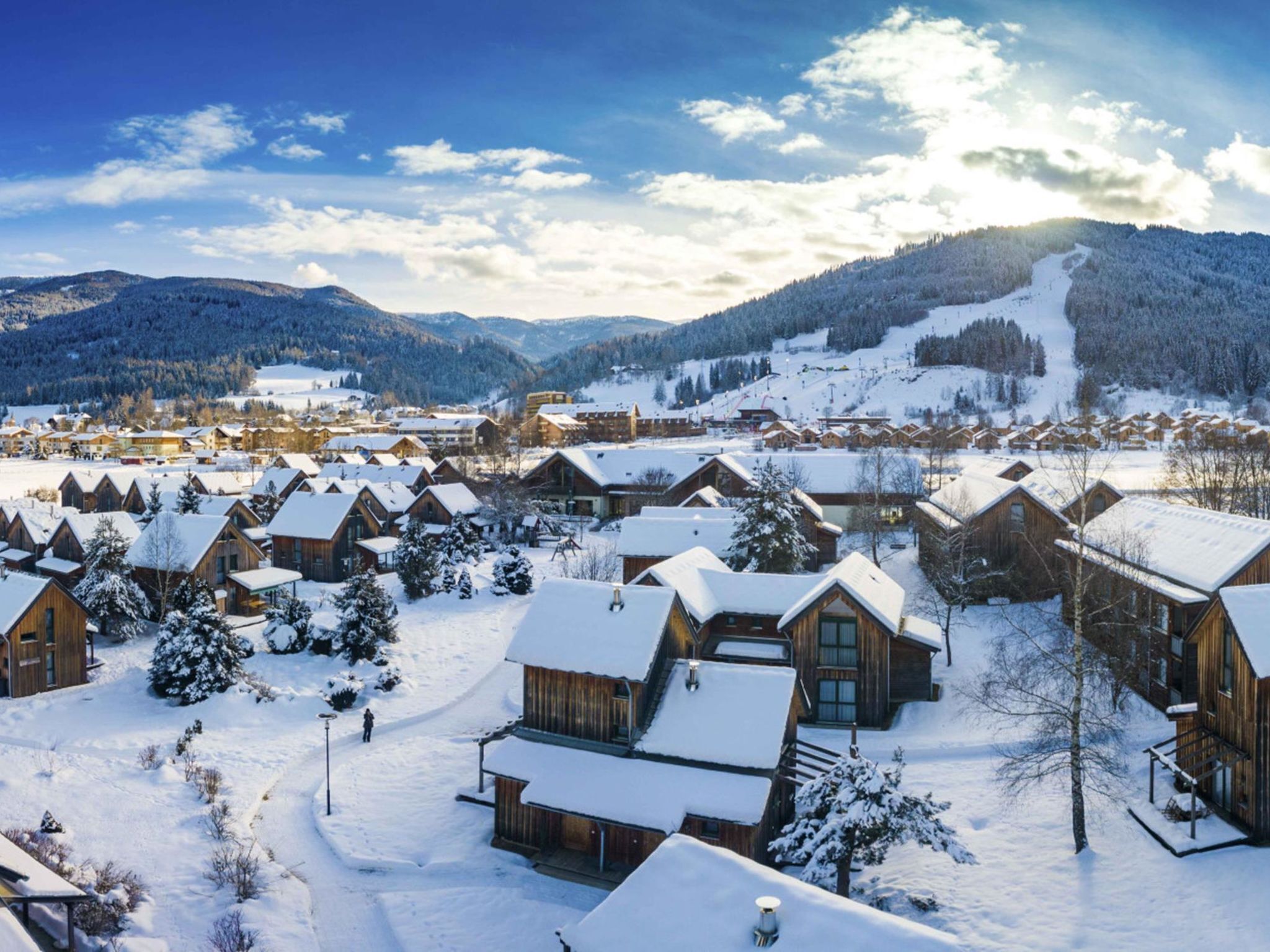 Un pueblo cubierto de nieve con cabañas de madera, una pista de esquí y montañas al fondo.