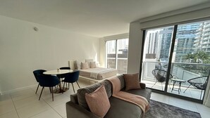 Una sala de estar moderna con un sofá, una mesa de comedor con sillas y un balcón con vistas a edificios altos.