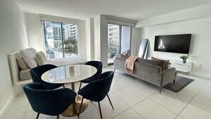 Una sala de estar moderna con una mesa de comedor, sillas, un sofá, un televisor y ventanas amplias con vistas a la ciudad.