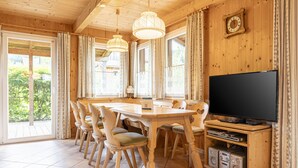 Comedor de madera con televisor y sistema de audio, ventanas amplias y vistas a la vegetación.