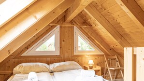 Un dormitorio con techo inclinado, dos ventanas, una cama, una lámpara de noche y una escalera.