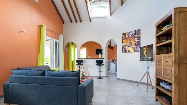 A living room with a sofa, a coffee table, a bookshelf, and a kitchen in the background. - Villa Ti'Twoine - Sea view - Sunsets - (Pointe-Noire)