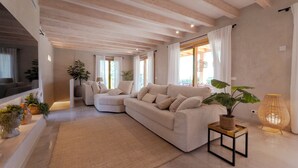 A modern living room with a beige sofa, a small side table with a plant, and a large window with curtains. - An oasis in the middle of nature (Pollença)
