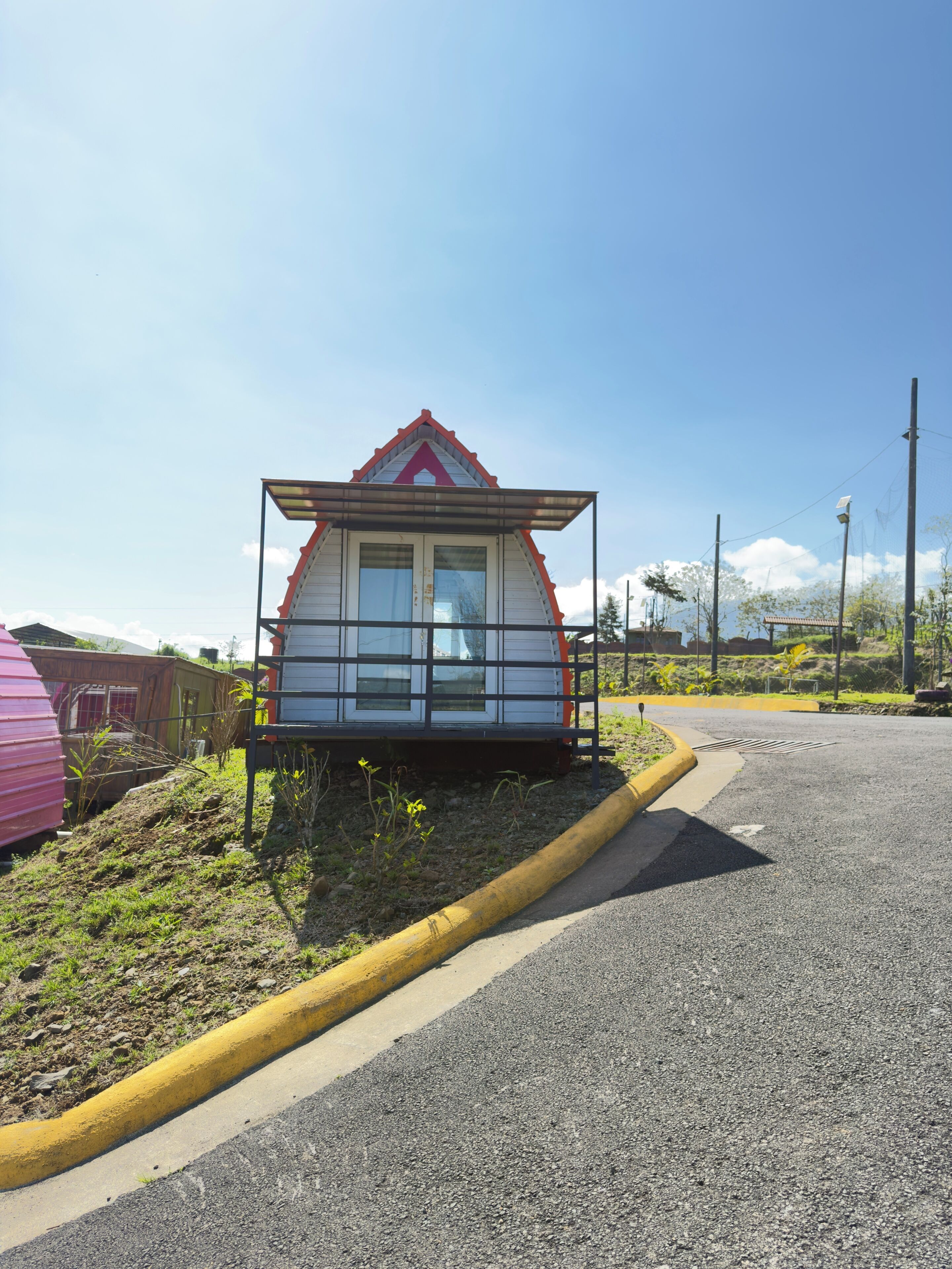 A small, single-story building with a red roof and white walls, situated on a grassy area beside a road.