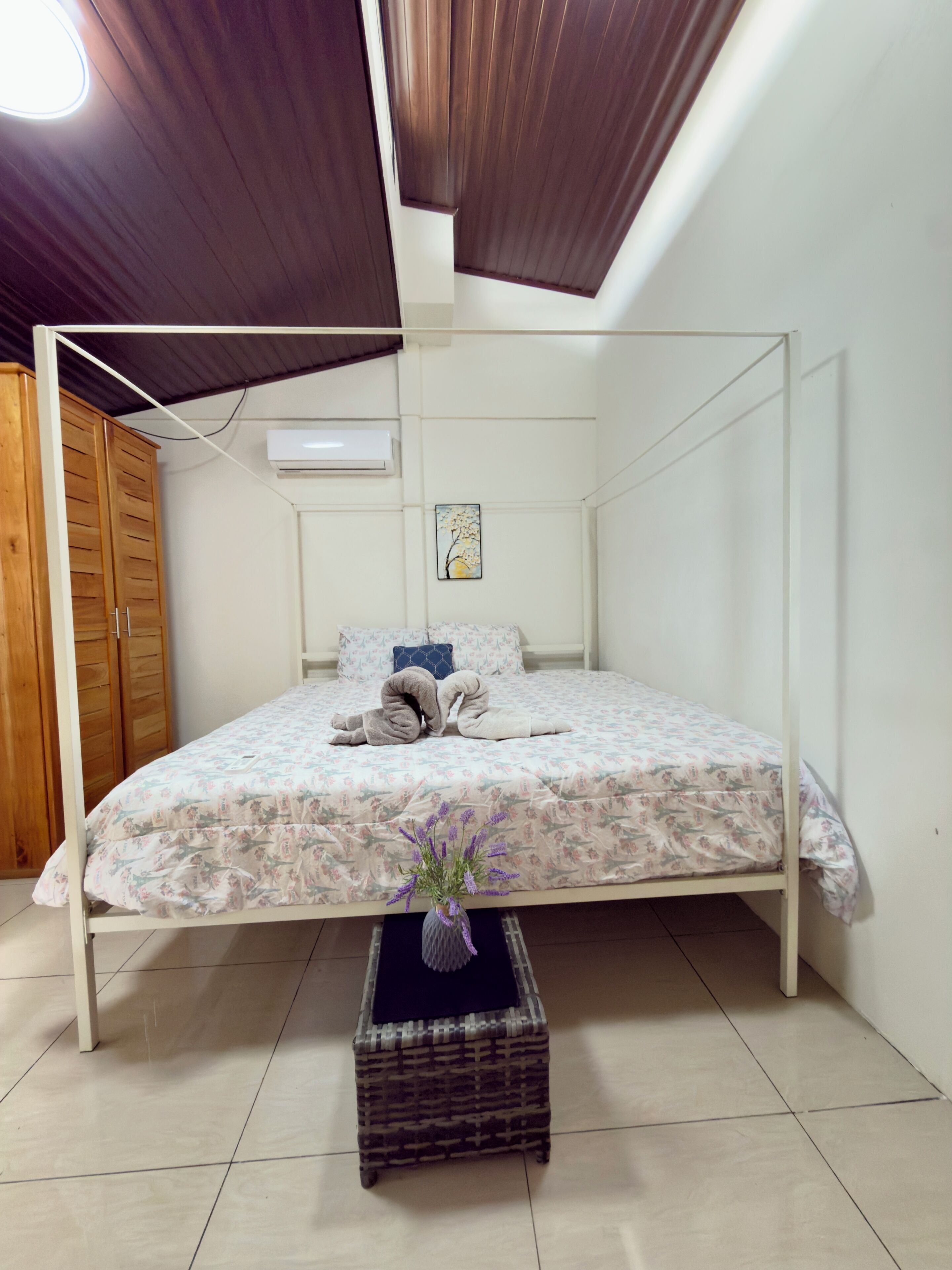 Un letto singolo con copriletto floreale, un comodino con un vaso di fiori, un condizionatore d'aria a muro e un armadio in legno.