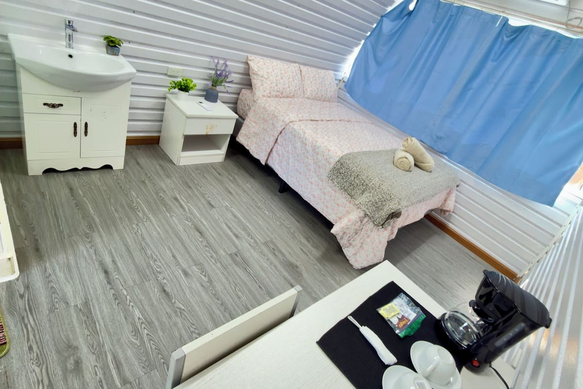 A compact, modern room with a bed, a small bedside table, a white sink, and a window with a blue curtain.