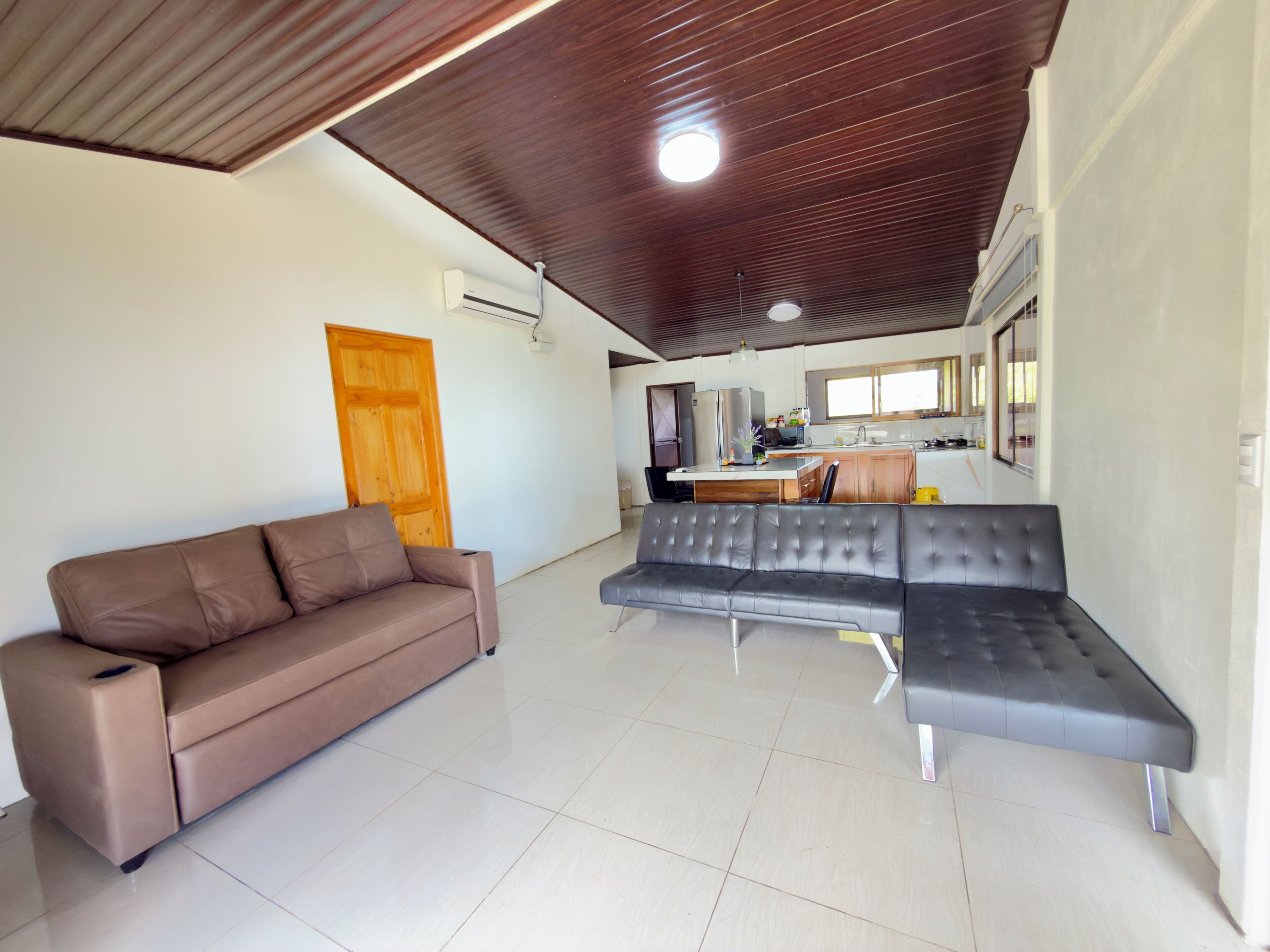 A living room with a brown sofa, a grey sectional sofa, a wooden ceiling, and a kitchen in the background.