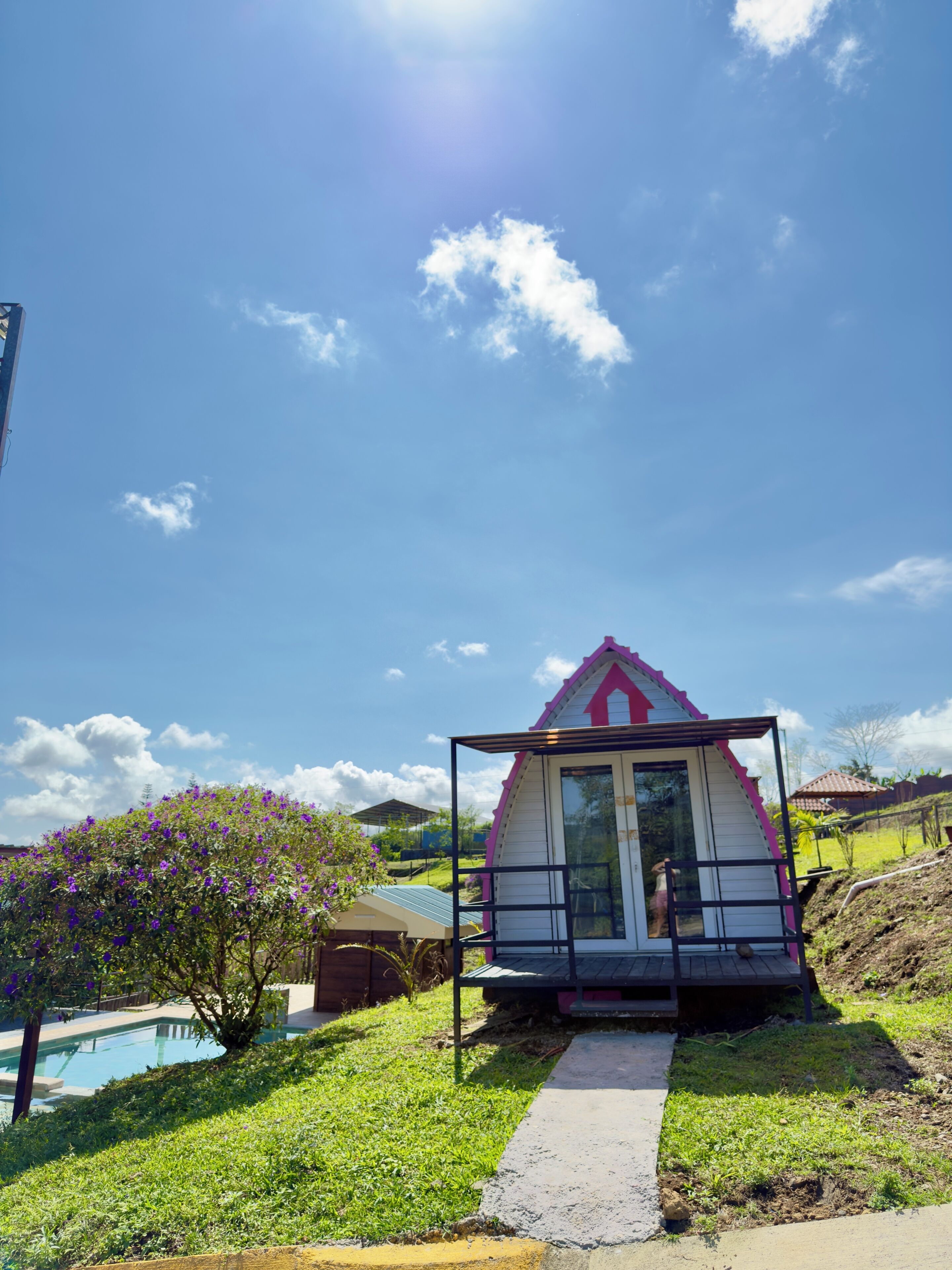 Una casita con techo rosa y puerta de cristal, rodeada de un jardín y una piscina.
