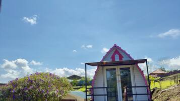 Una casita con techo rosa y puerta de cristal, rodeada de un jardín y una piscina.