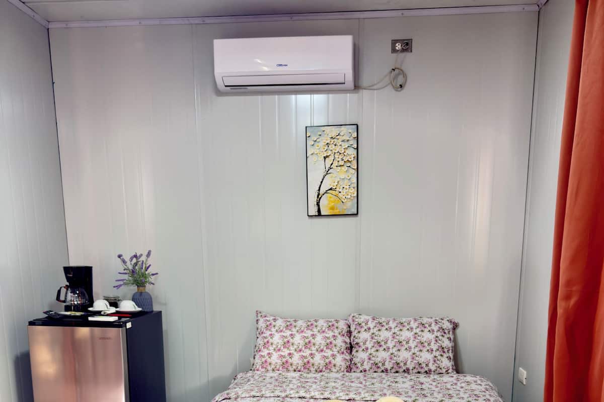 A bedroom with a bed, a small desk with a coffee maker, an air conditioner, and a painting on the wall.