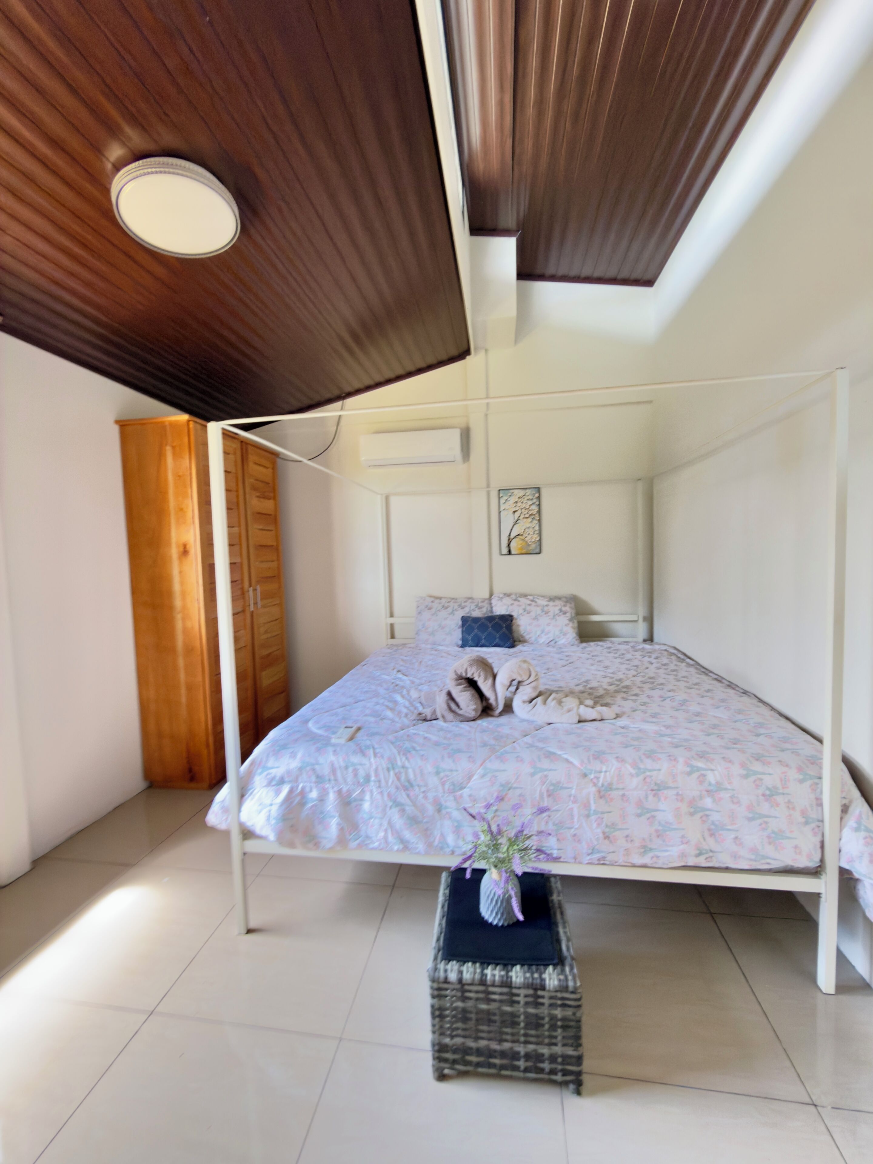 Une chambre à coucher avec un lit, une armoire en bois, un petit tabouret en osier et un ventilateur de plafond.