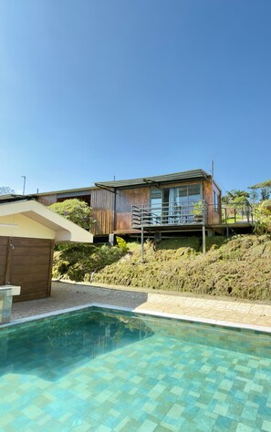 Een modern huis met een zwembad, een houten terras en uitzicht op het omliggende landschap.
